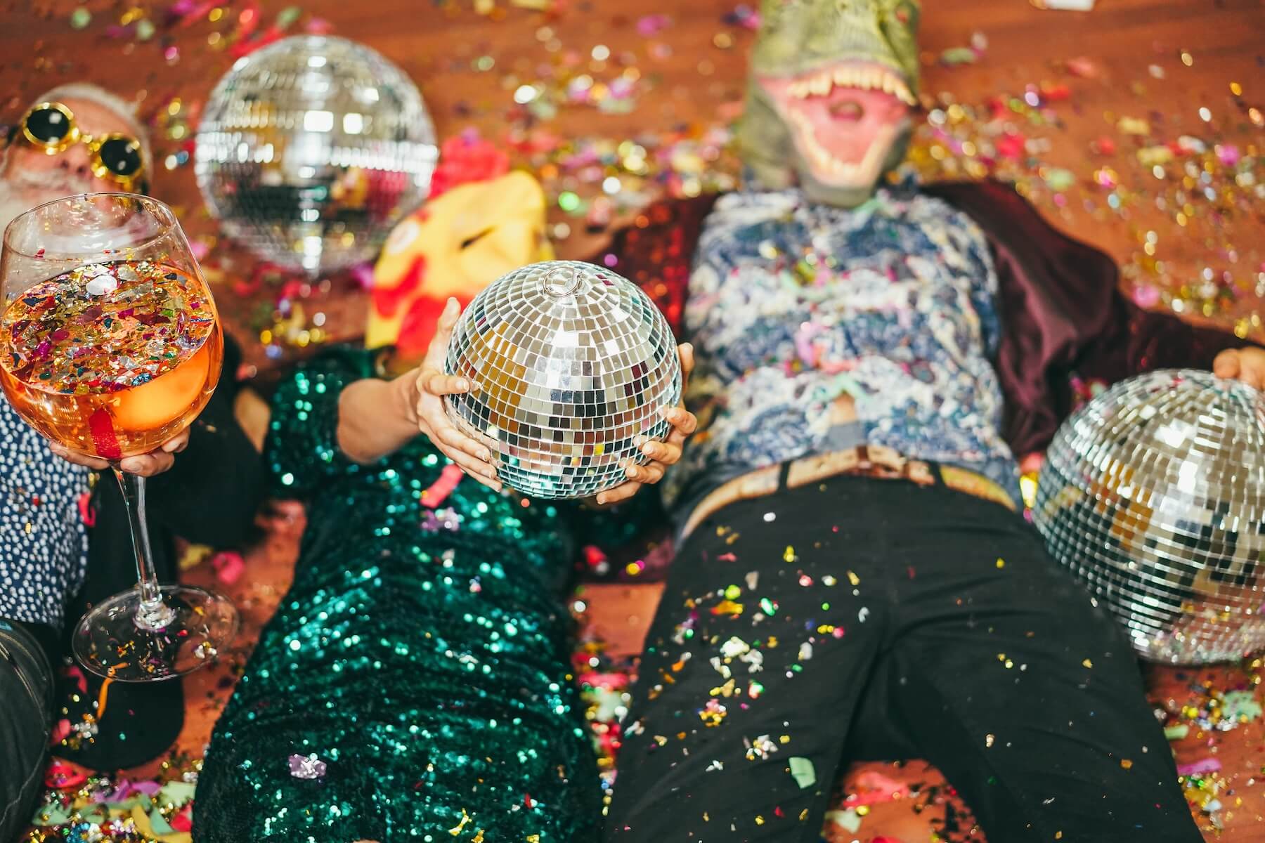 Three people at a party lying on the floor with disco balls, confetti scattered, one person wearing a dinosaur mask.