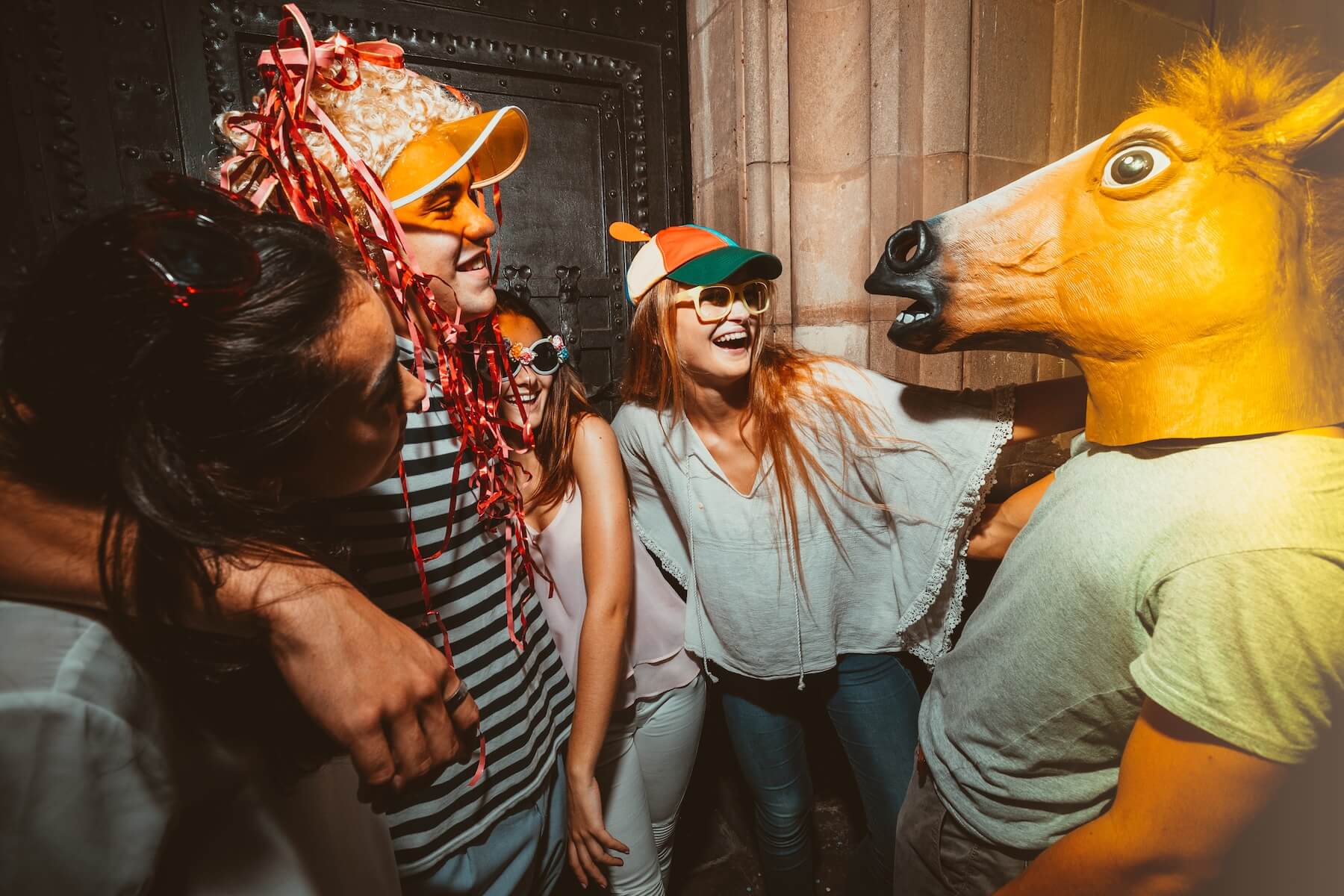 Group of four friends laughing in costume — one wearing a horse mask and others in silly hats.